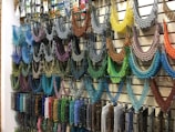 A wall display featuring an array of intricately designed beaded necklaces in a variety of colors hanging from metal hooks. Each necklace is crafted with multicolored beads arranged in symmetrical, semi-circular patterns, offering a vivid and artistic presentation. The setup conveys a sense of vibrant artistry typical in hand-crafted jewelry shops.