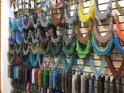 A wall display featuring an array of intricately designed beaded necklaces in a variety of colors hanging from metal hooks. Each necklace is crafted with multicolored beads arranged in symmetrical, semi-circular patterns, offering a vivid and artistic presentation. The setup conveys a sense of vibrant artistry typical in hand-crafted jewelry shops.