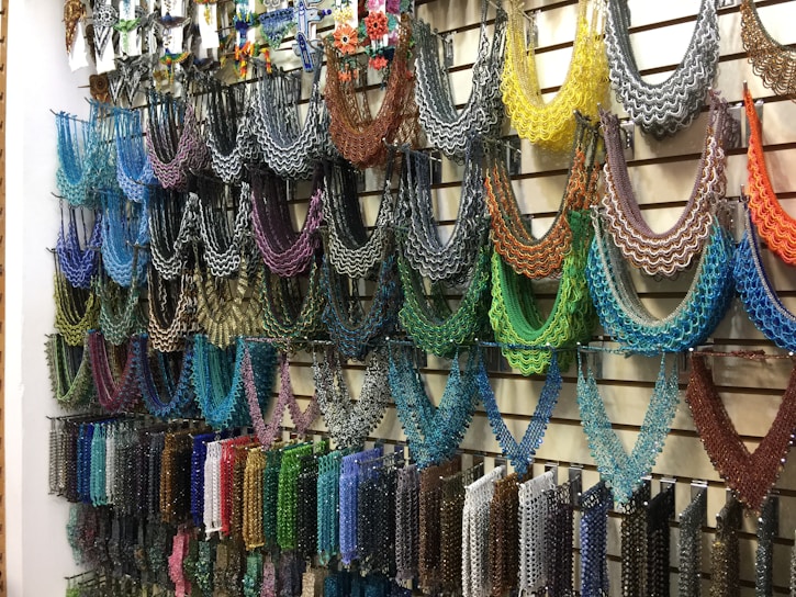 A wall display featuring an array of intricately designed beaded necklaces in a variety of colors hanging from metal hooks. Each necklace is crafted with multicolored beads arranged in symmetrical, semi-circular patterns, offering a vivid and artistic presentation. The setup conveys a sense of vibrant artistry typical in hand-crafted jewelry shops.