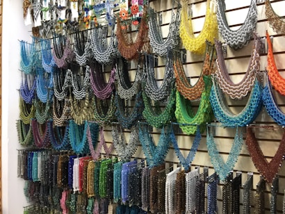 A wall display featuring an array of intricately designed beaded necklaces in a variety of colors hanging from metal hooks. Each necklace is crafted with multicolored beads arranged in symmetrical, semi-circular patterns, offering a vivid and artistic presentation. The setup conveys a sense of vibrant artistry typical in hand-crafted jewelry shops.