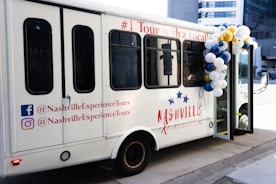 A white touring bus with the text 'Nashville Experience Tours' displayed on its side. The bus is decorated with balloons in white, blue, and gold. Social media handles are printed on the bus's exterior, including logos for Facebook and Instagram. The bus has multiple dark-tinted windows and is parked on a city street.