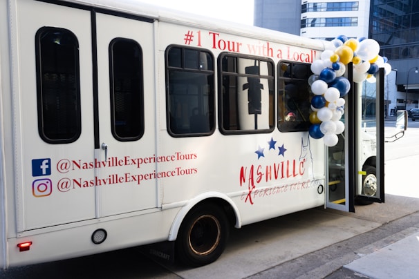 A white touring bus with the text 'Nashville Experience Tours' displayed on its side. The bus is decorated with balloons in white, blue, and gold. Social media handles are printed on the bus's exterior, including logos for Facebook and Instagram. The bus has multiple dark-tinted windows and is parked on a city street.