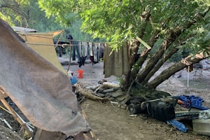 A makeshift campsite in a wooded area featuring a tent constructed from tarps and cloth, with clothing hung to dry on a line between the trees. Various items, including a black plastic bag and a stroller, are scattered around the site. The ground is covered with dry leaves and the area is shaded by green trees.