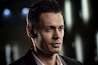 A man with short, dark hair and a clean-shaven face is wearing a dark suit and white shirt. The background is blurred with bokeh lights, giving a dramatic and focused look to the portrait.