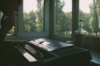 A person is sitting by a large window, playing a keyboard in a dimly lit room. The person has a cigarette in their mouth. A bottle and a glass are placed on the windowsill, with trees visible through the window outside.