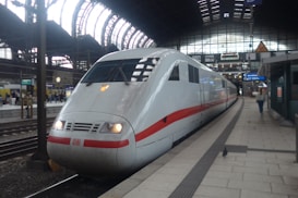 A modern white and red high-speed train is stationed at a railway platform inside a large, ornate train station. The structure features expansive arched windows, and there are passengers visible in the distance on another platform. The train showcases a sleek design, with a pointed nose and large windows.