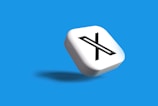 A white 3D cube with a black letter 'X' prominently displayed on one face, set against a vibrant blue background. The cube casts a subtle shadow, giving a sense of depth and dimension.