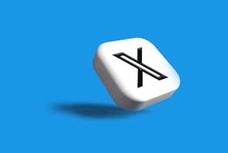 A white 3D cube with a black letter 'X' prominently displayed on one face, set against a vibrant blue background. The cube casts a subtle shadow, giving a sense of depth and dimension.