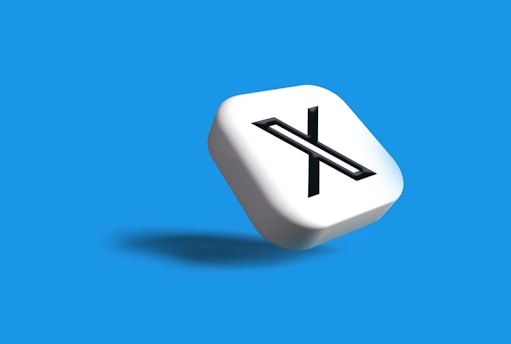 A white 3D cube with a black letter 'X' prominently displayed on one face, set against a vibrant blue background. The cube casts a subtle shadow, giving a sense of depth and dimension.