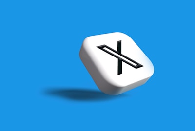 A white 3D cube with a black letter 'X' prominently displayed on one face, set against a vibrant blue background. The cube casts a subtle shadow, giving a sense of depth and dimension.