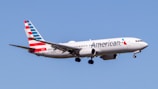 An airplane with American Airlines livery is flying in a clear blue sky. The aircraft is a narrow-body jet with a distinct red, white, and blue pattern on its tail.