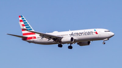 An airplane with American Airlines livery is flying in a clear blue sky. The aircraft is a narrow-body jet with a distinct red, white, and blue pattern on its tail.
