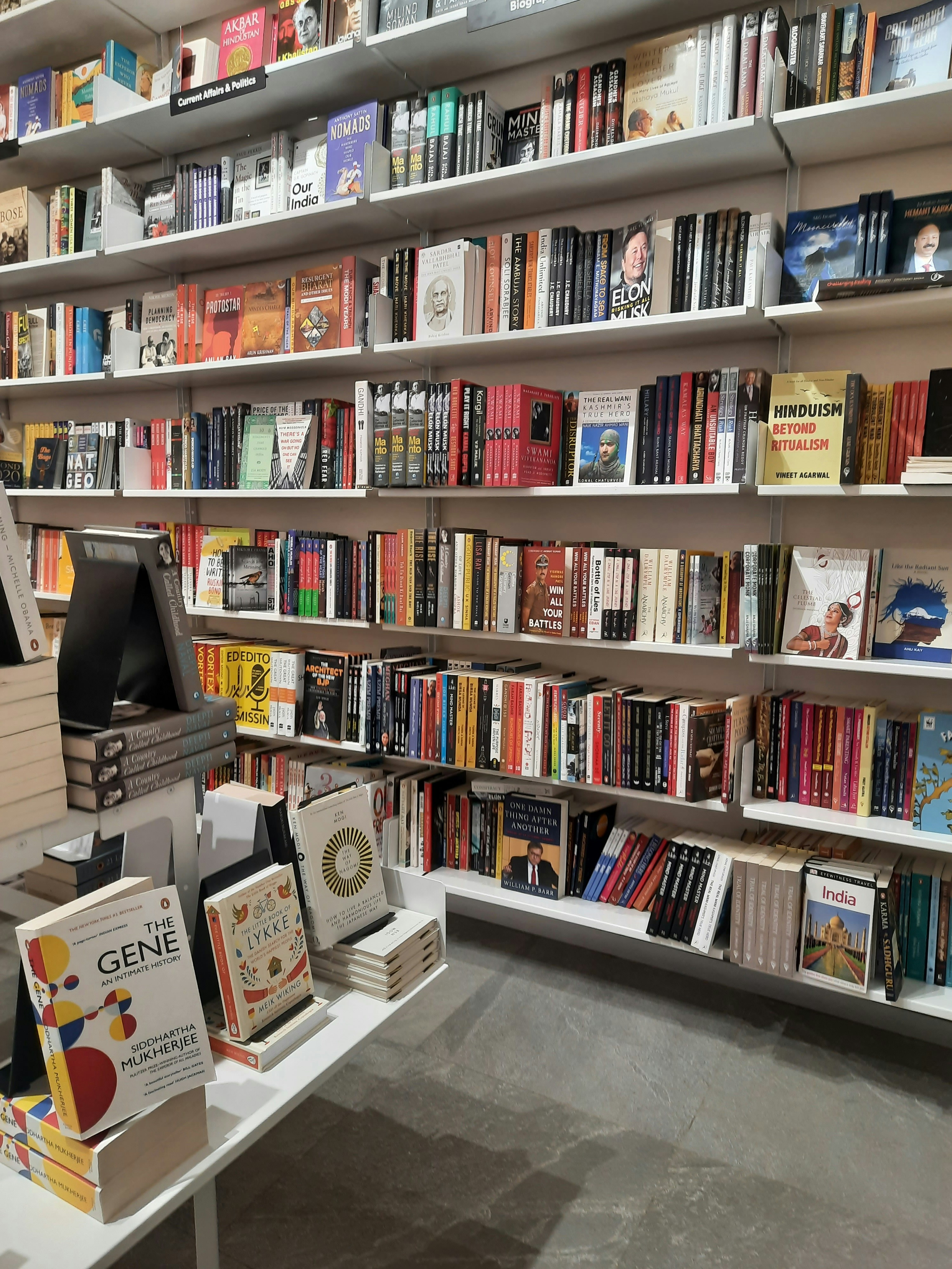 Bookshelves filled with a variety of books are organized neatly in a bookstore. The shelves hold an assortment of genres, including politics, science, and culture. A display table in the foreground showcases a small selection of highlighted titles.
