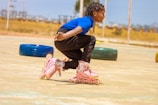 A child wearing a blue top and black pants is skating on a pair of pink rollerblades. They are captured in a crouched position, suggesting movement or a skating maneuver. The background includes blurred elements like a tire, indicating an outdoor or recreational environment.