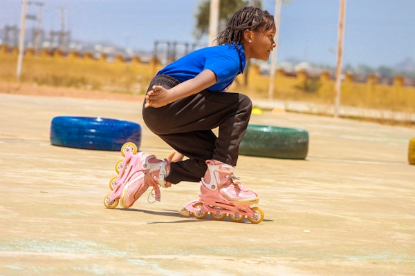 A child wearing a blue top and black pants is skating on a pair of pink rollerblades. They are captured in a crouched position, suggesting movement or a skating maneuver. The background includes blurred elements like a tire, indicating an outdoor or recreational environment.