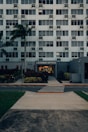A modern multi-story building with numerous windows, featuring a central entrance labeled 'Leasing Office'. A pathway leads up to the entrance, bordered by manicured plants and palm trees. A zebra crossing stretches across the asphalt road in front of the building.