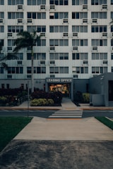 A modern multi-story building with numerous windows, featuring a central entrance labeled 'Leasing Office'. A pathway leads up to the entrance, bordered by manicured plants and palm trees. A zebra crossing stretches across the asphalt road in front of the building.
