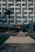 A modern multi-story building with numerous windows, featuring a central entrance labeled 'Leasing Office'. A pathway leads up to the entrance, bordered by manicured plants and palm trees. A zebra crossing stretches across the asphalt road in front of the building.