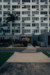 A modern multi-story building with numerous windows, featuring a central entrance labeled 'Leasing Office'. A pathway leads up to the entrance, bordered by manicured plants and palm trees. A zebra crossing stretches across the asphalt road in front of the building.