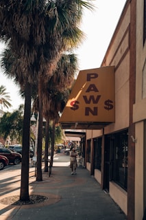 A sidewalk lined with palm trees and a yellow sign indicating a pawn shop. The street features parked cars and a person walking. The scene is well-lit with natural light, and the atmosphere is calm.