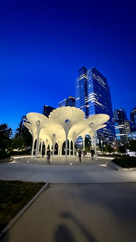 A modern architectural canopy structure illuminated at night with a backdrop of tall, glass-covered skyscrapers against a deep blue sky. People are walking around the well-lit area surrounded by greenery.