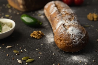 A loaf of rustic bread with a dusting of flour rests on a dark surface, surrounded by assorted seeds, a piece of walnut, a green chili, and a small bowl of cream cheese. In the background, there are other bread pieces and tomatoes.