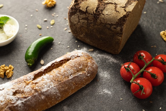 A rustic arrangement of a loaf of bread and a baguette, accompanied by a cluster of red cherry tomatoes, a green chili pepper, walnuts, and a small dish of cream cheese garnished with mint. The scene is set on a dark textured surface sprinkled with seeds, contributing to an artisanal and natural aesthetic.