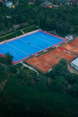 An aerial view of a large sports complex with a vibrant blue field surrounded by a red boundary and adjacent clay courts. The complex is nestled amidst dense greenery and trees, with a few buildings visible in the background. The setting appears to be serene and well-maintained, with a sense of spaciousness.