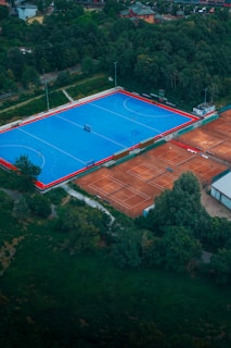 An aerial view of a large sports complex with a vibrant blue field surrounded by a red boundary and adjacent clay courts. The complex is nestled amidst dense greenery and trees, with a few buildings visible in the background. The setting appears to be serene and well-maintained, with a sense of spaciousness.