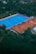 An aerial view of a large sports complex with a vibrant blue field surrounded by a red boundary and adjacent clay courts. The complex is nestled amidst dense greenery and trees, with a few buildings visible in the background. The setting appears to be serene and well-maintained, with a sense of spaciousness.