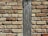 A close-up of a brick wall with a vertical wooden beam running through the center. The bricks are of varying sizes and shades of brown, beige, and tan, with visible mortar between them. The wooden beam has a rough texture and weathered appearance, contrasting with the uniformity of the bricks.