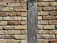 A close-up of a brick wall with a vertical wooden beam running through the center. The bricks are of varying sizes and shades of brown, beige, and tan, with visible mortar between them. The wooden beam has a rough texture and weathered appearance, contrasting with the uniformity of the bricks.