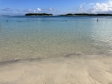 Clear blue waters gently lap against a sandy beach under a bright blue sky, with distant green islands visible on the horizon. A line of buoys floats on the water, marking a boundary.