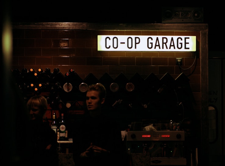A dark indoor setting with a sign reading 'CO-OP GARAGE' illuminated above a counter. The background includes a rack of various wine bottles and glasses. Two people are partially visible in the foreground under low lighting conditions, likely standing behind or near the counter.