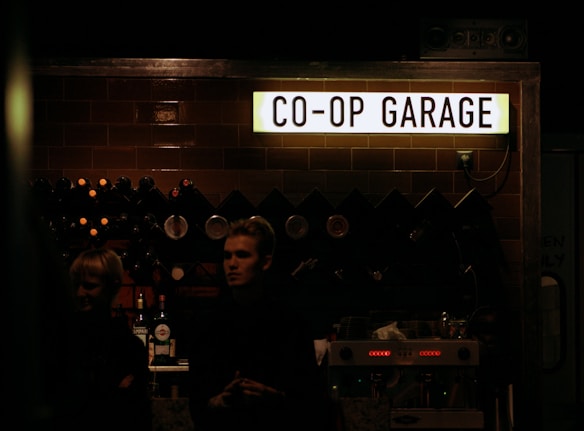 A dark indoor setting with a sign reading 'CO-OP GARAGE' illuminated above a counter. The background includes a rack of various wine bottles and glasses. Two people are partially visible in the foreground under low lighting conditions, likely standing behind or near the counter.