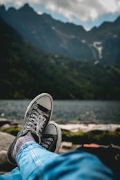 Casual sneakers with black canvas and white rubber soles are worn by someone who is relaxing by a serene lake. The background features a view of distant mountains under a cloudy sky.