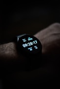 A close-up view of a smartwatch displaying the time, date, battery percentage, heart rate, and step count on its digital screen. The watch is worn on a wrist, and the background is dark, emphasizing the illuminated display.