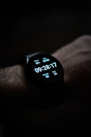 A close-up view of a smartwatch displaying the time, date, battery percentage, heart rate, and step count on its digital screen. The watch is worn on a wrist, and the background is dark, emphasizing the illuminated display.