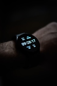 A close-up view of a smartwatch displaying the time, date, battery percentage, heart rate, and step count on its digital screen. The watch is worn on a wrist, and the background is dark, emphasizing the illuminated display.