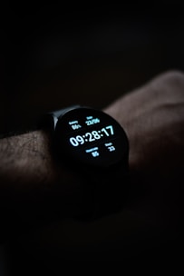 A close-up view of a smartwatch displaying the time, date, battery percentage, heart rate, and step count on its digital screen. The watch is worn on a wrist, and the background is dark, emphasizing the illuminated display.
