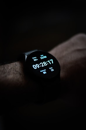 A close-up view of a smartwatch displaying the time, date, battery percentage, heart rate, and step count on its digital screen. The watch is worn on a wrist, and the background is dark, emphasizing the illuminated display.