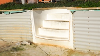 An empty swimming pool is shown with visible steps leading out. The pool walls have a corrugated metal texture, and signs of rust and water stains are evident. The surroundings include patches of grass at the top edge and some scattered debris on the pool floor.