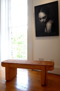 A wooden bench with a tiled surface is positioned on a polished wooden floor in a brightly lit room. Natural light pours in through tall windows on the left. On the wall, there is a large framed black-and-white photograph of a person with a serious expression, the details are slightly blurred, creating a somber or mysterious effect.