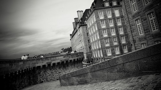 A grayscale image featuring historic stone buildings with multiple windows lining a street. The architecture displays classic European design elements. Several people are gathered along a stone wall, which runs parallel to the buildings. The sky appears overcast, adding a dramatic effect to the scene.