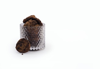 A glass jar with a diamond pattern holds multiple chocolate chip cookies, with one cookie resting against the jar on a plain white surface.