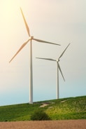 Two large wind turbines stand prominently on a grassy hill, surrounded by lush greenery and under a clear sky with soft lighting.