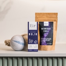 Several wellness products are arranged on a surface. There is a box labeled 'Eir Health Sleep No. 10' that indicates it contains CBD products for calming and sleep aid, featuring evening oil with lavender and melatonin. Beside it, a pouch labeled 'Midnight Chill' contains bath salts. In front, there is a bath bomb with a subtle gray and blue design.