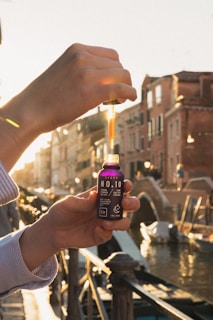 A hand holding a bottle with a dropper dispensing liquid against a picturesque canal with rustic buildings in the background. The sunlight casts a warm glow, highlighting the text 'NO.10' and 'Eir' on the bottle, indicating it is a CBD oil product.