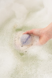 A hand is holding a bath bomb, partially submerged in soapy bathwater. Bubbles and foam surround the area, with a slight tint of green in the water.