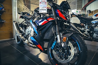 A high-performance motorcycle is prominently displayed in a showroom. It features sleek, aerodynamic design elements with a mix of blue, red, and black colors. The motorcycle is positioned on a glossy, dark-tiled floor, and a price tag is attached above the handlebar. Additional motorcycles and a helmet are visible in the background, along with a poster on the wall.